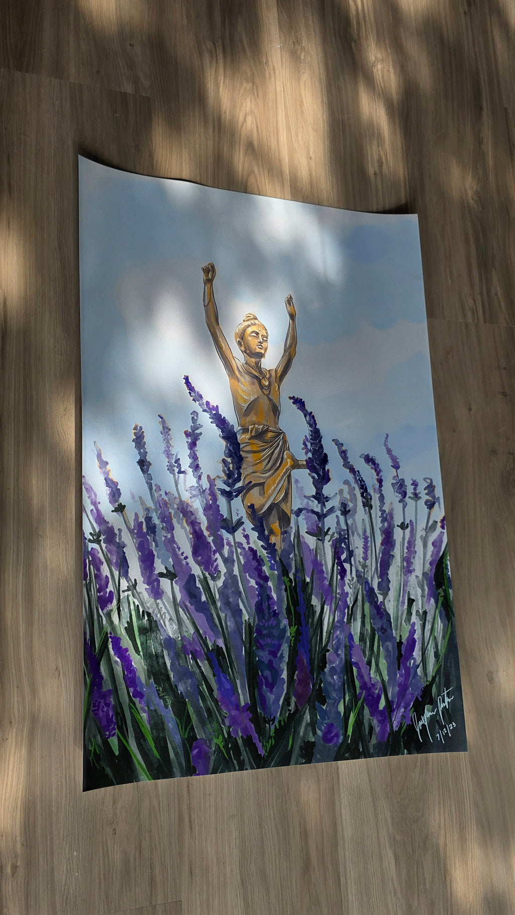 A vertical wall tapestry featuring a painted scene of a golden statue among purple lavender fields under a cloudy sky, displayed hanging on a wooden floor with dappled sunlight.