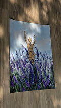 A vertical wall tapestry featuring a painted scene of a golden statue among purple lavender fields under a cloudy sky, displayed hanging on a wooden floor with dappled sunlight.
