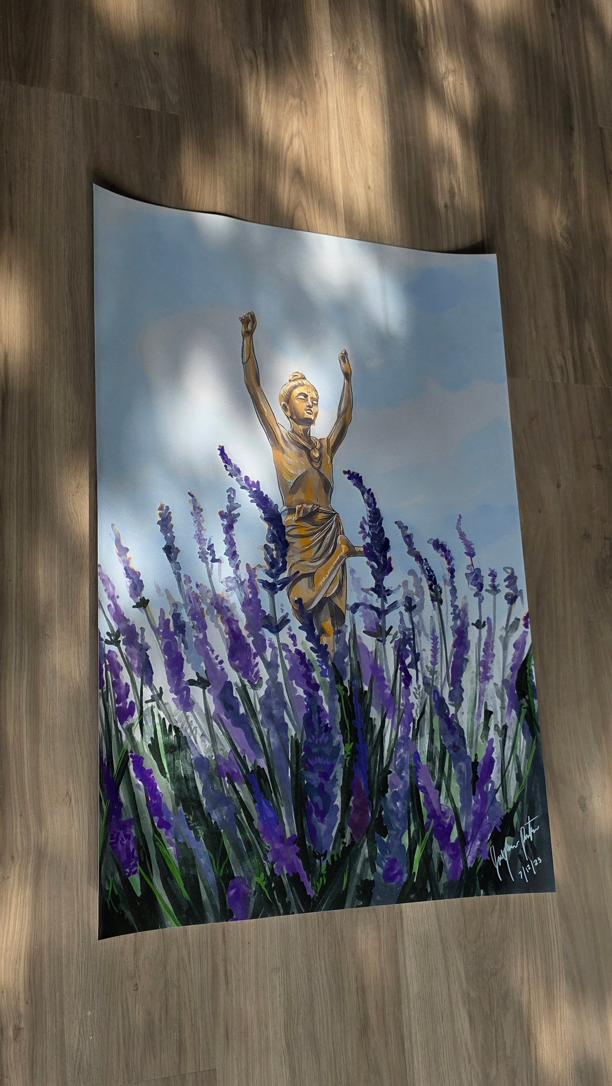 A vertical wall tapestry featuring a painted scene of a golden statue among purple lavender fields under a cloudy sky, displayed hanging on a wooden floor with dappled sunlight.