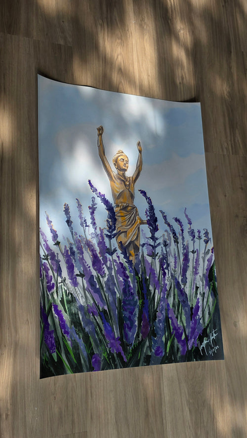 A vertical wall tapestry featuring a painted scene of a golden statue among purple lavender fields under a cloudy sky, displayed hanging on a wooden floor with dappled sunlight.